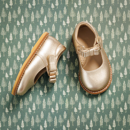 Toddler girls' metallic gold Mary Jane shoes with bow detail and tan rubber soles on green patterned background.