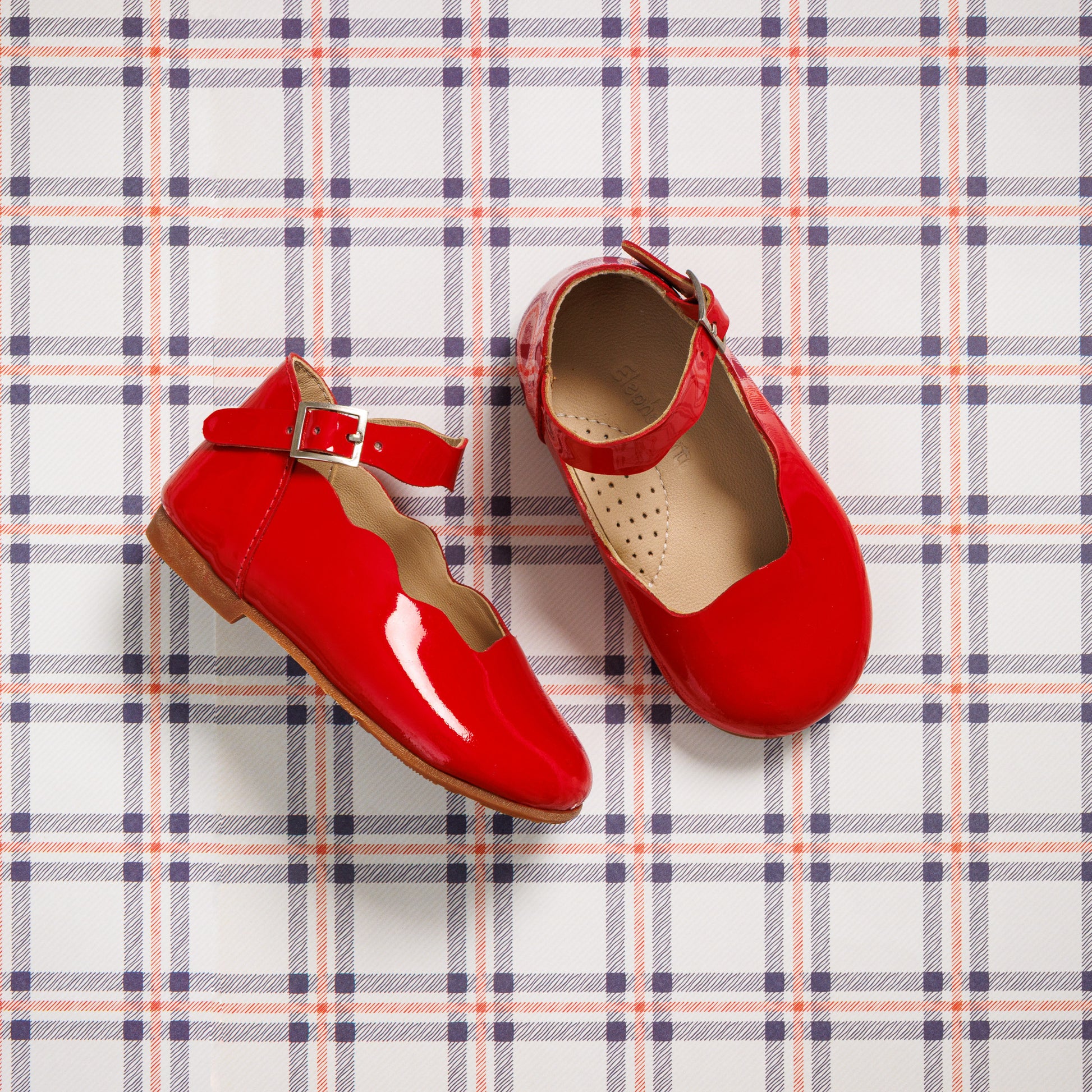 Glossy red toddler flats with ankle straps and scalloped edges on plaid background.