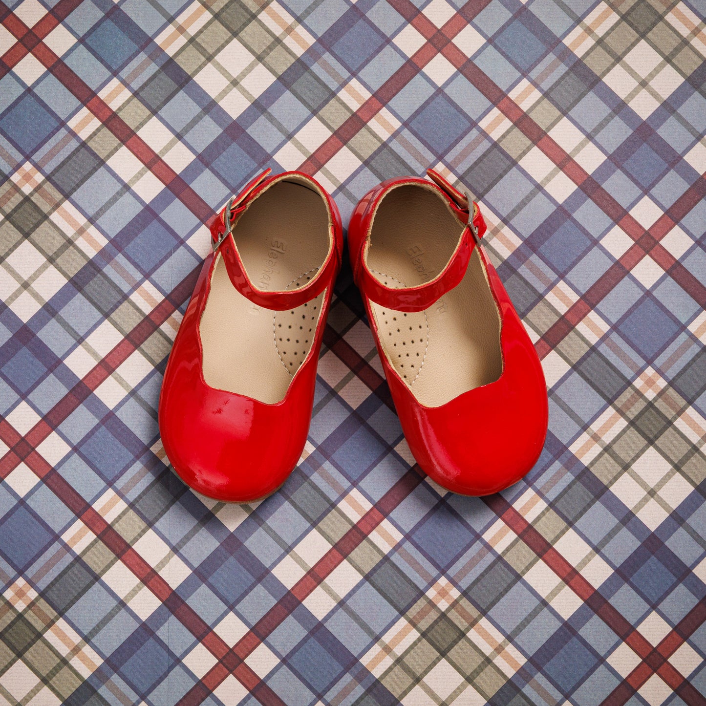 Glossy red toddler Mary Jane flats with ankle straps on a checkered blue, white, and red background.