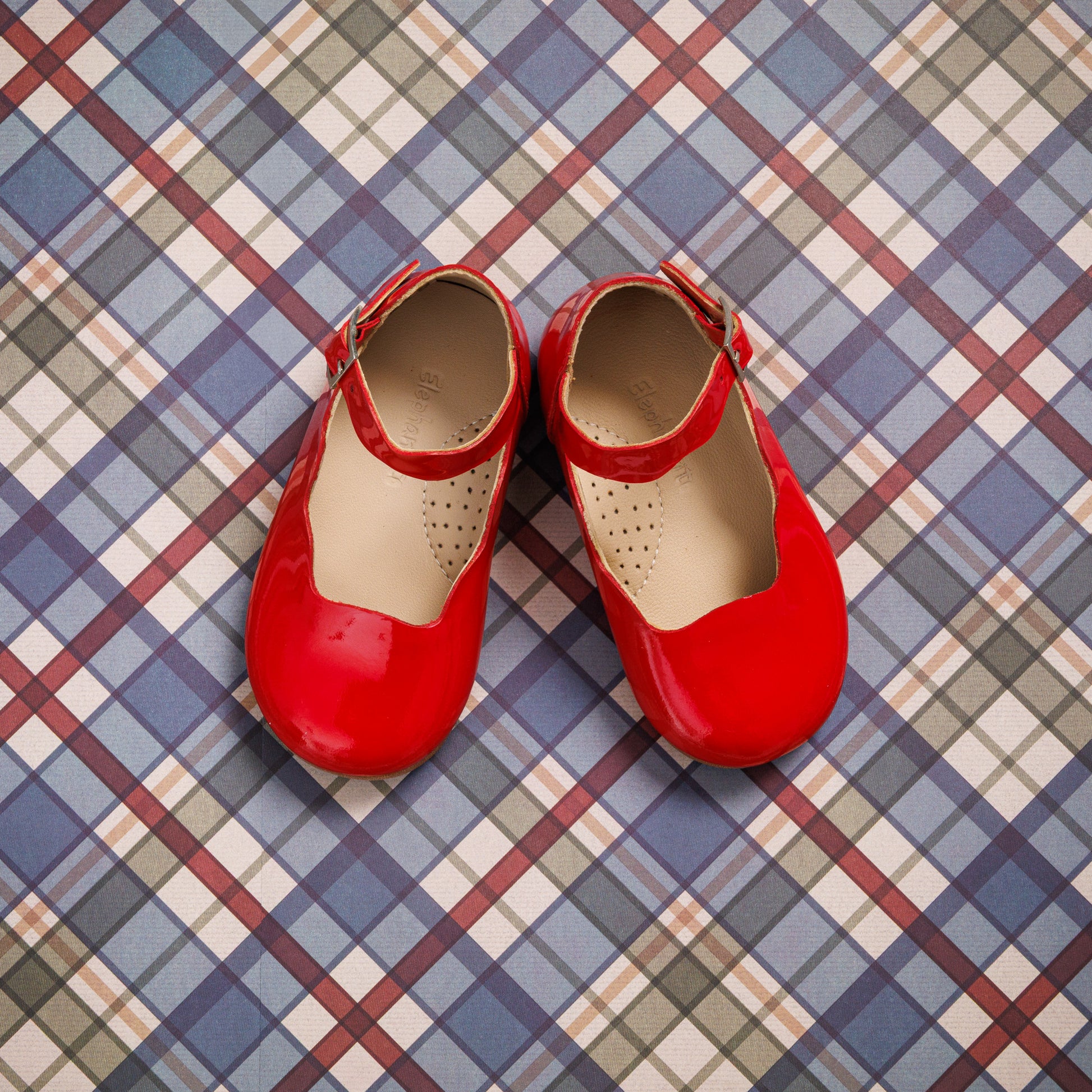 Glossy red toddler Mary Jane flats with ankle straps on a checkered blue, white, and red background.