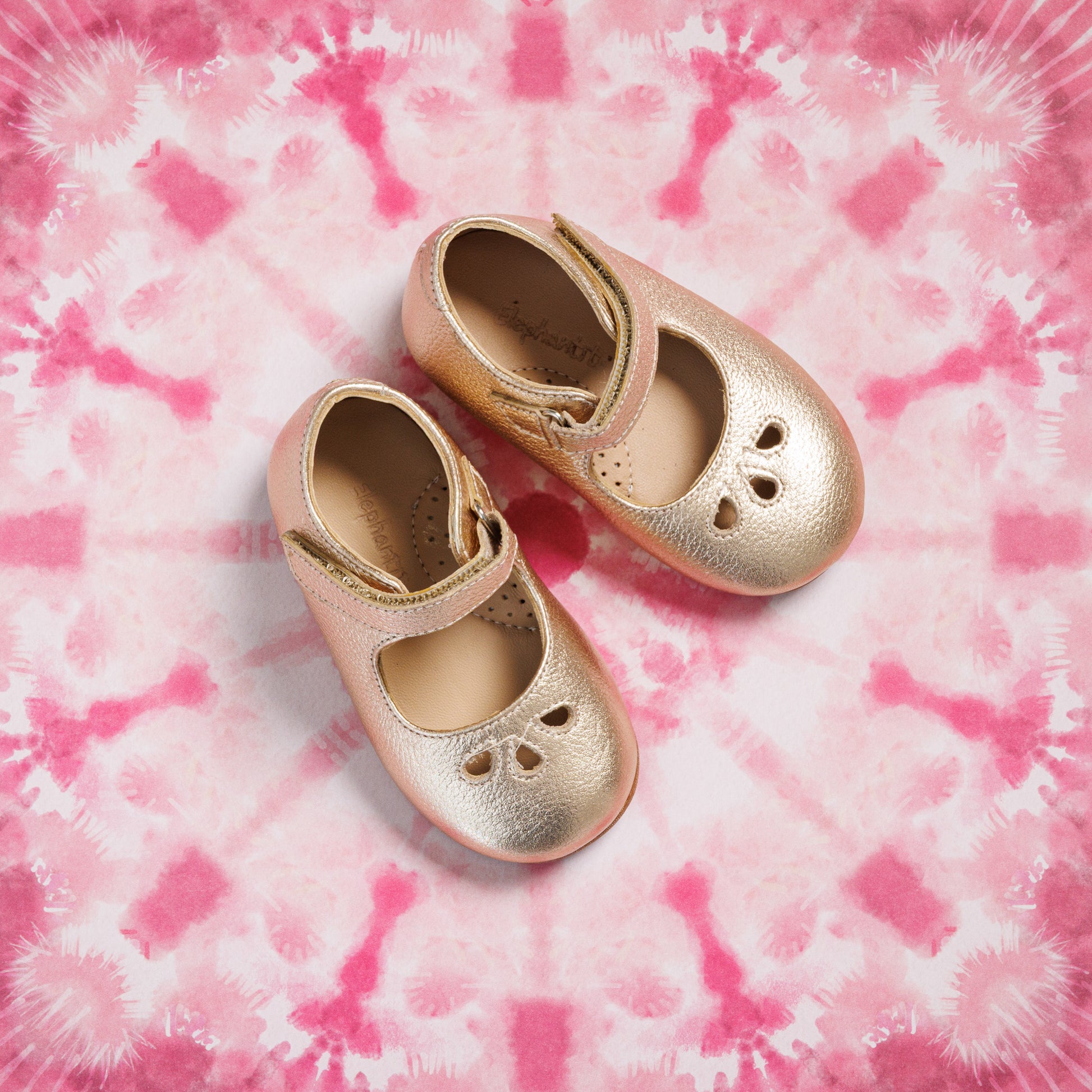 Toddler gold Mary Jane shoes with heart cutouts and adjustable strap on pink tie-dye background.