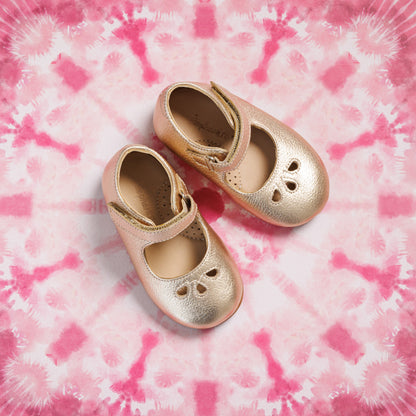 Toddler gold Mary Jane shoes with heart cutouts and adjustable strap on pink tie-dye background.