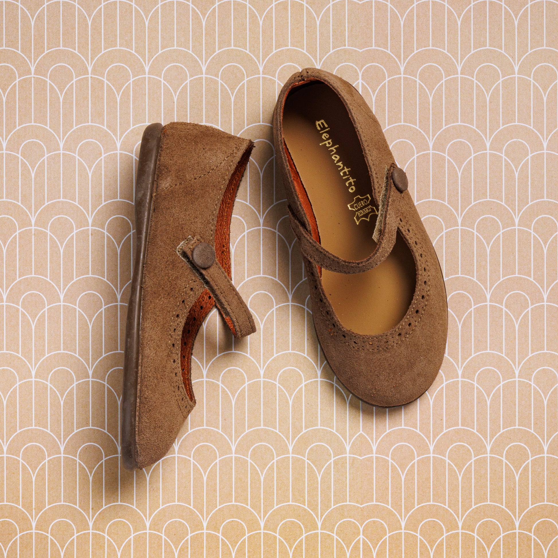 A pair of Elephantito Suede Mary Jane Beige shoes for girls, with one flat and one on its side, shown on a tan geometric patterned background.