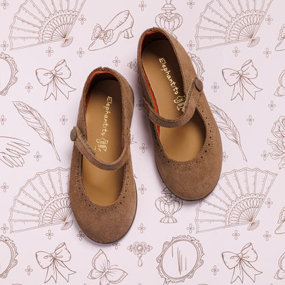 A pair of Suede Mary Jane Beige shoes by Elephantito with buttoned straps is displayed on paper featuring illustrated fans, bows, mirrors, feathers, and perfume bottles.
