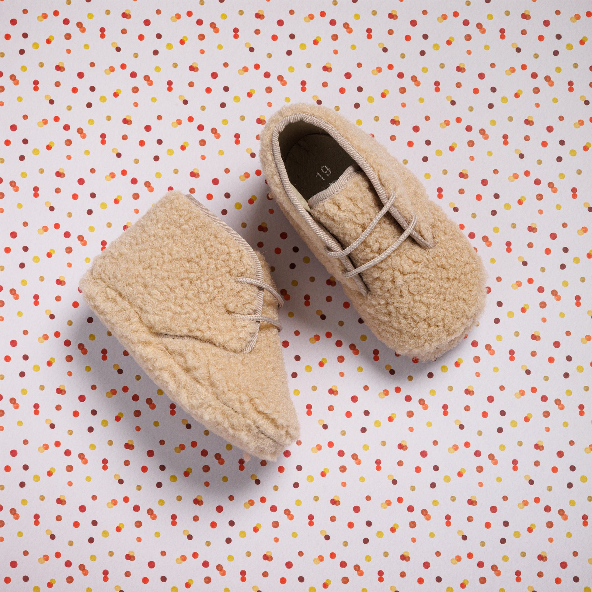 Beige shearling baby booties with soft texture and lace-up detail on multicolor polka dot background.