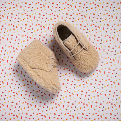 Beige shearling baby booties with soft texture and lace-up detail on multicolor polka dot background.