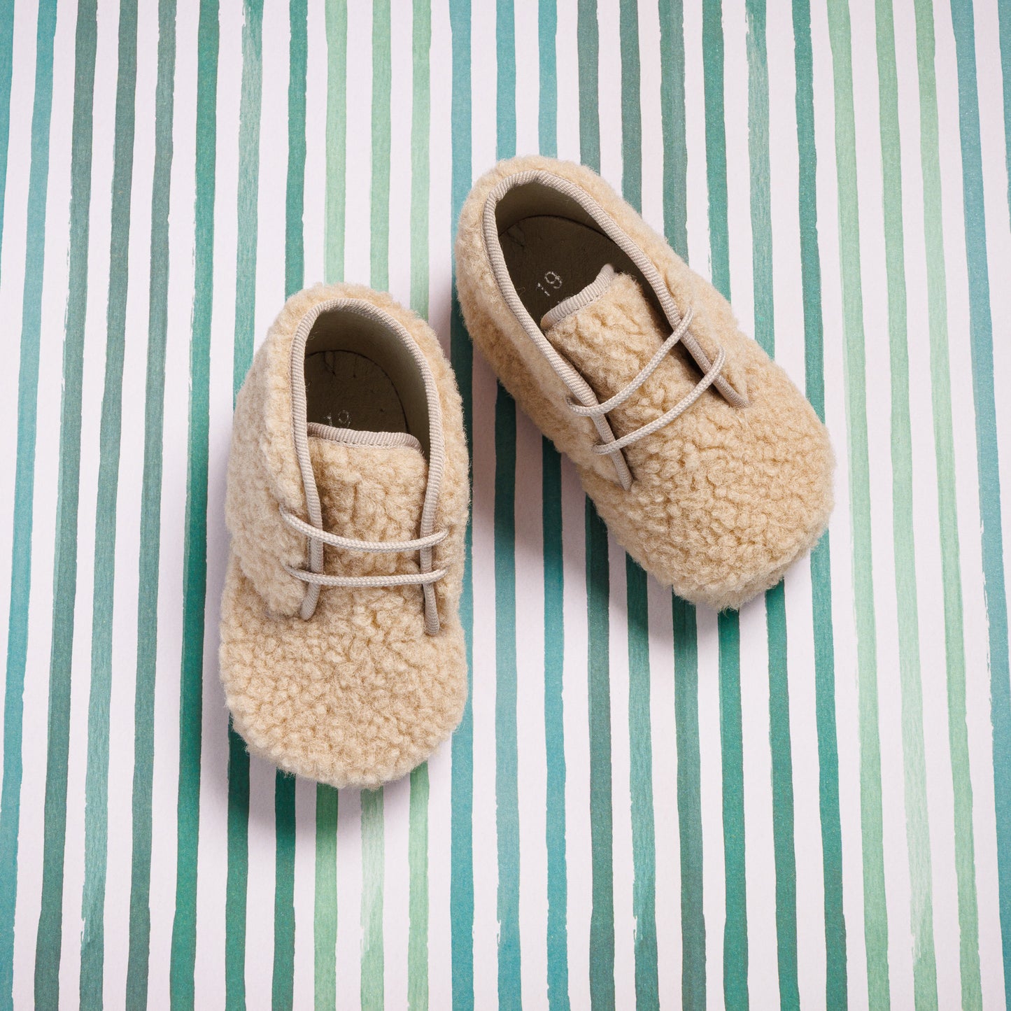 Beige shearling baby booties with laces on striped fabric background.