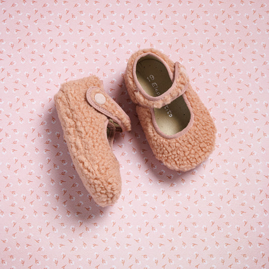 Pink shearling baby Mary Jane shoes with button strap and soft sole on pink patterned background.