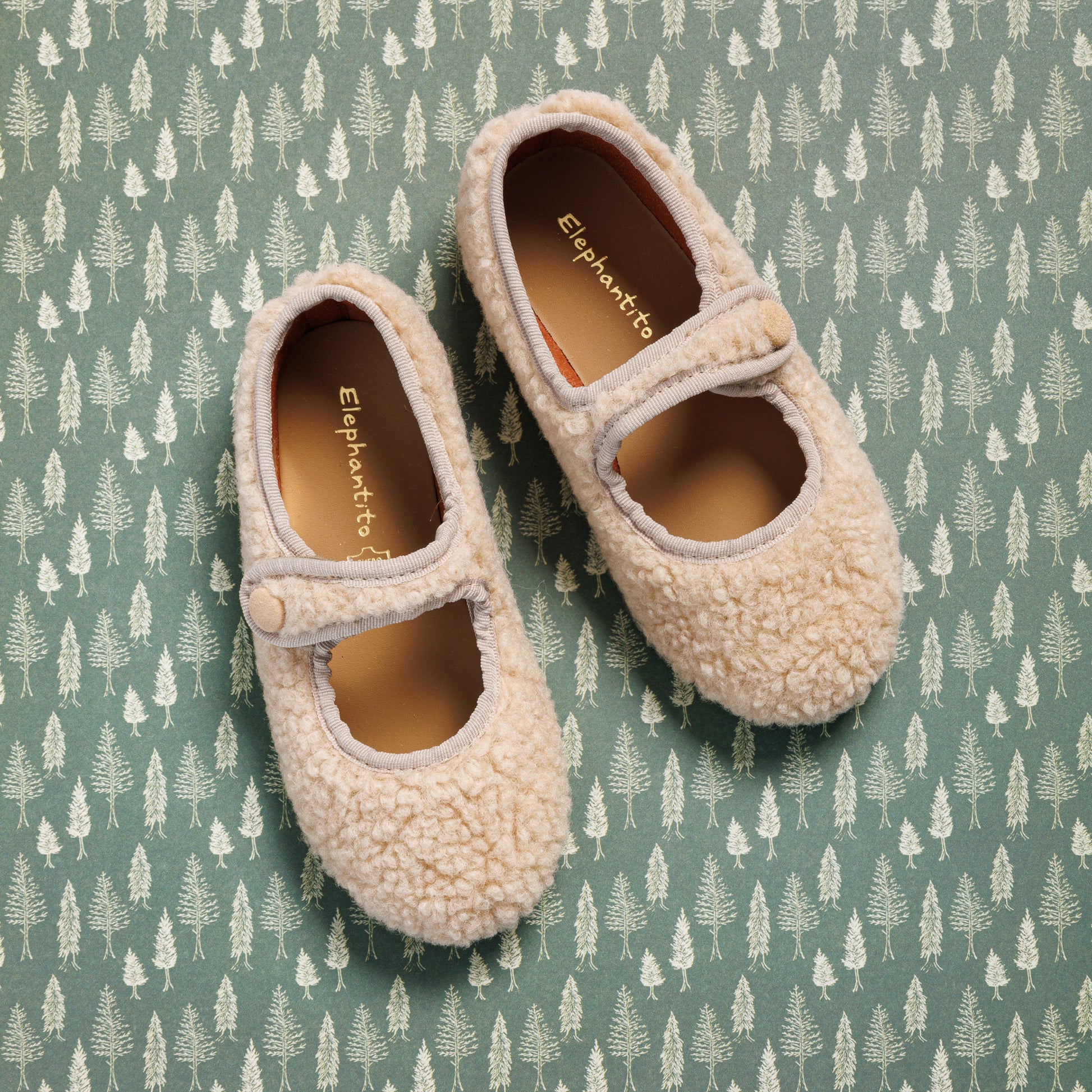 Beige shearling Mary Jane shoes with strap and button detail for toddlers on green patterned background.