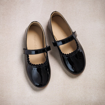 Black patent leather Mary Jane flats with scalloped edges and adjustable straps for kids.