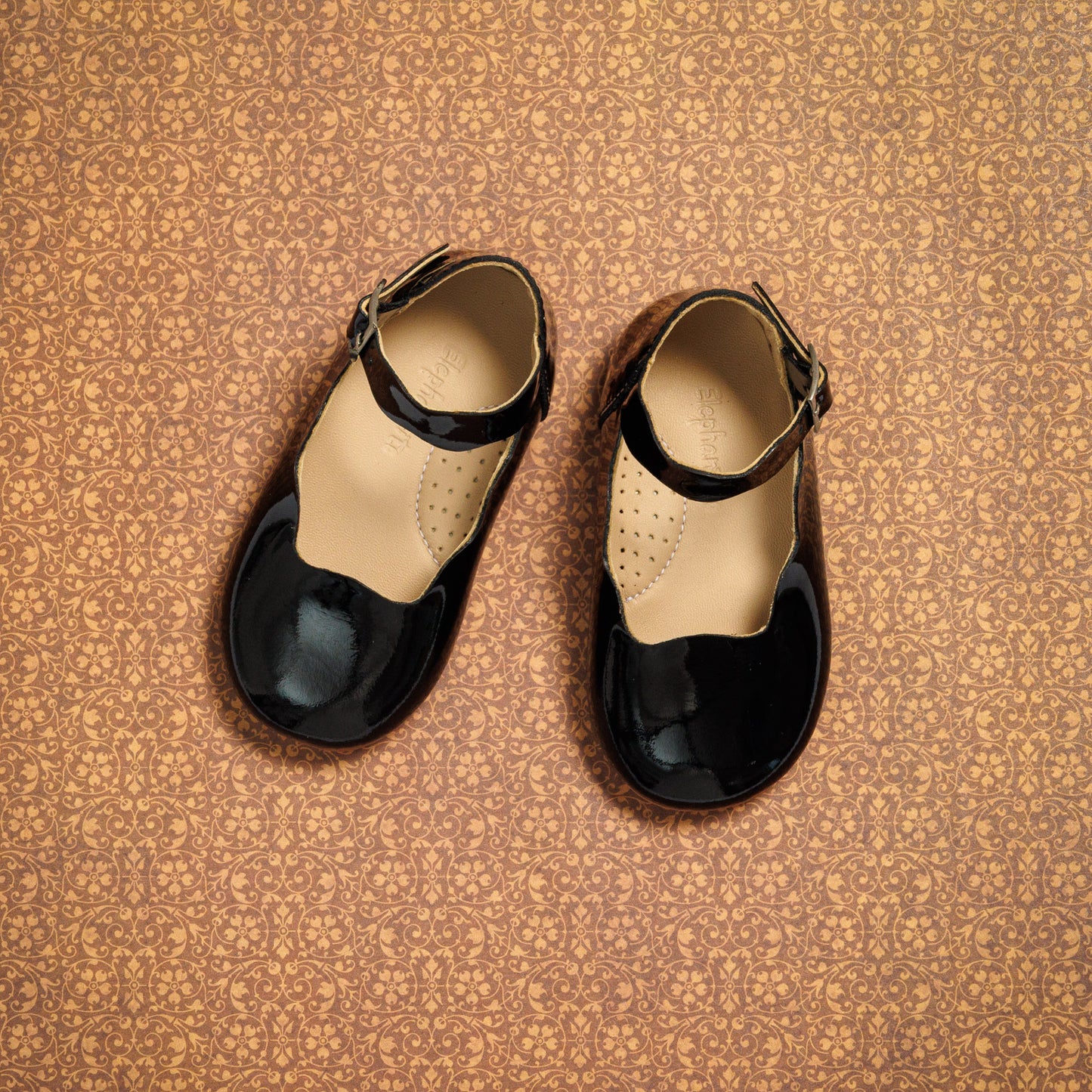 Black patent leather toddler flats with ankle strap and round toe on patterned brown surface.