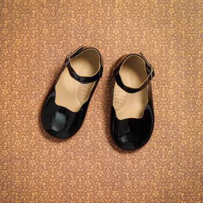 Black patent leather toddler flats with ankle strap and round toe on patterned brown surface.