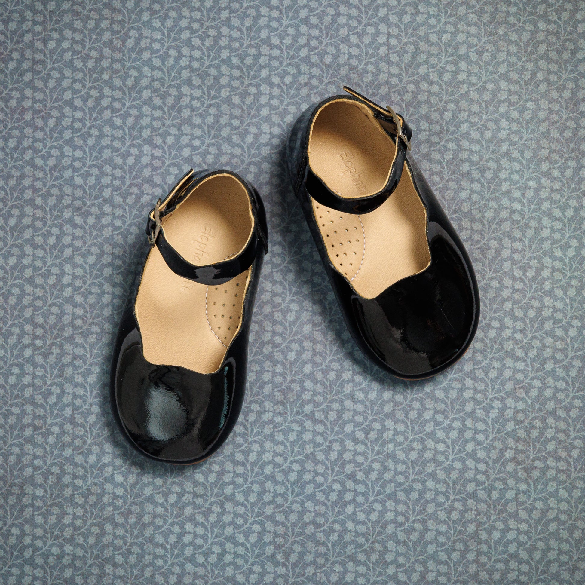 Black patent leather toddler flats with ankle straps and beige cushioned insoles on patterned gray background.