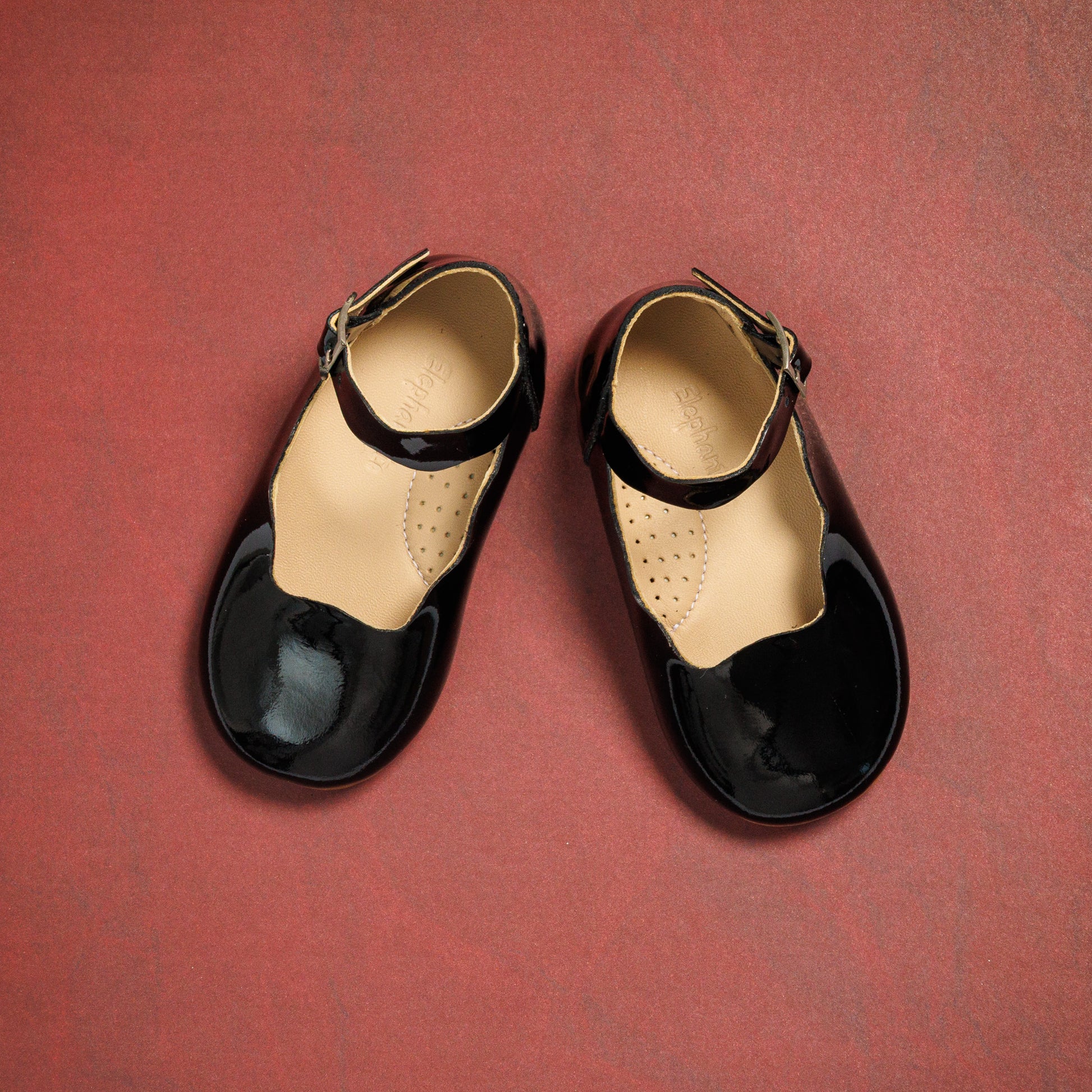 Black patent leather toddler flats with ankle straps and rounded toes on a red background.