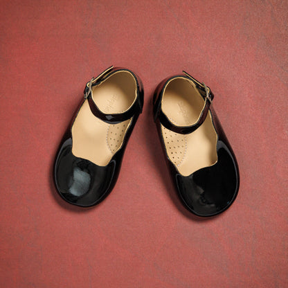 Black patent leather toddler flats with ankle straps and rounded toes on a red background.
