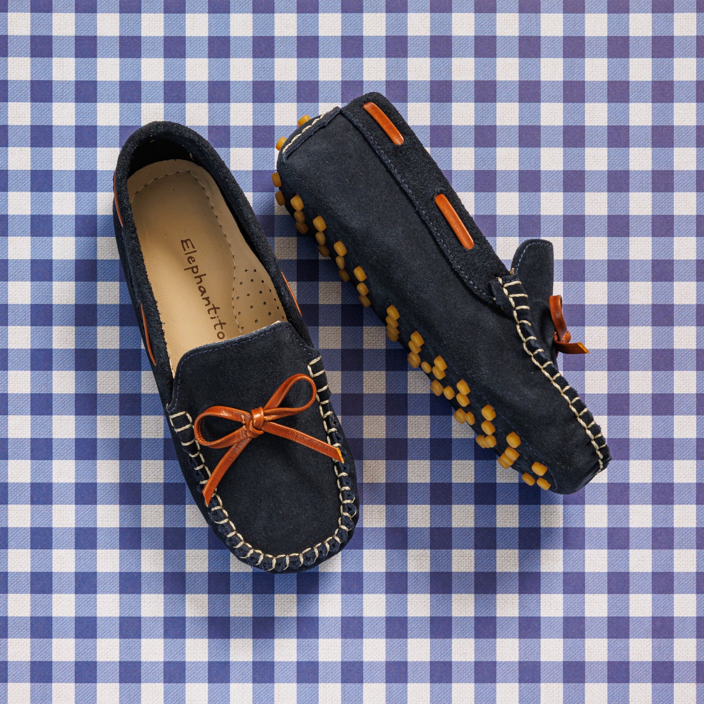 Elephantito - Navy blue suede boys' driver loafers with orange bow and rubber pebble sole detailing on gingham background.