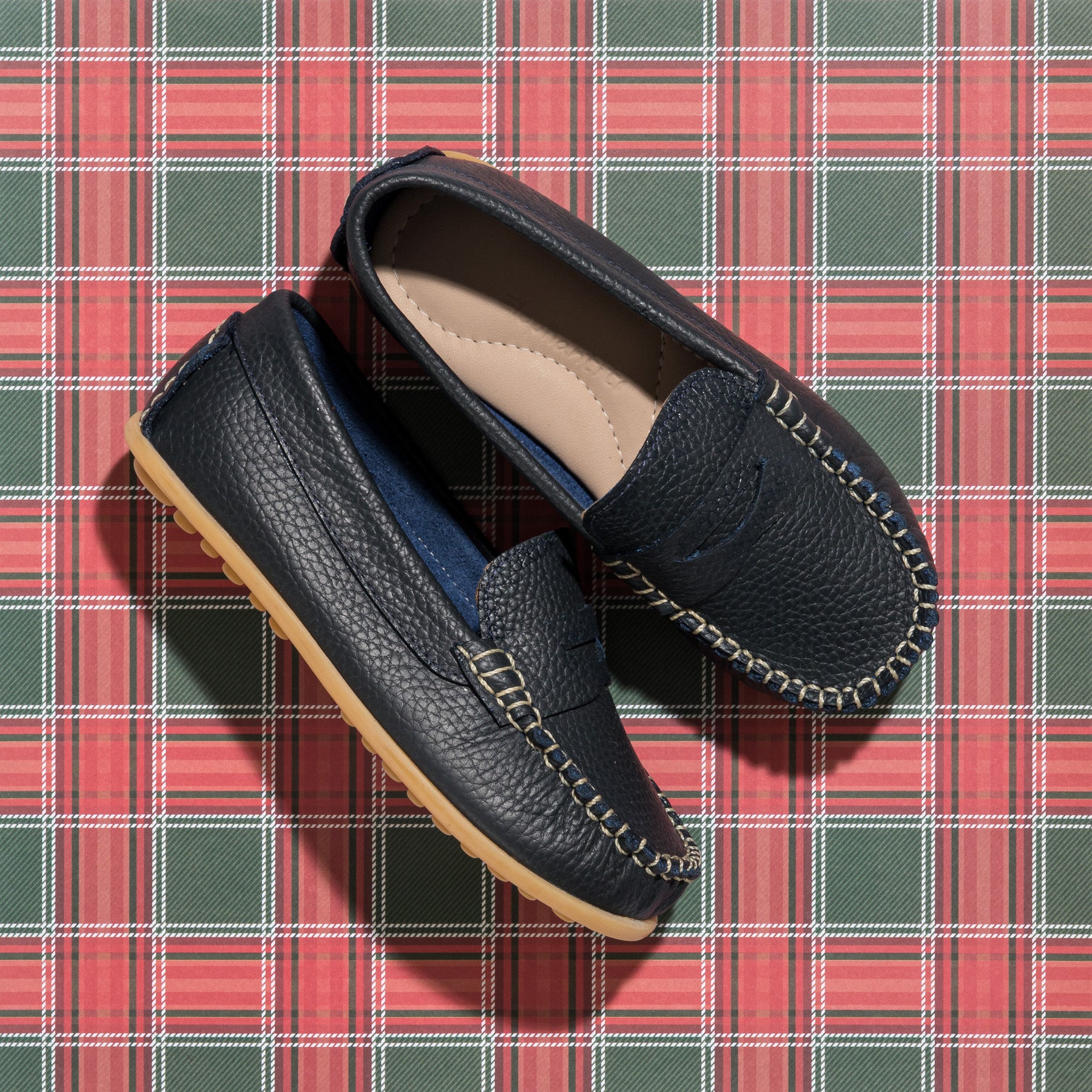 Navy blue leather boys loafers with tan stitching and rubber sole on red plaid background.