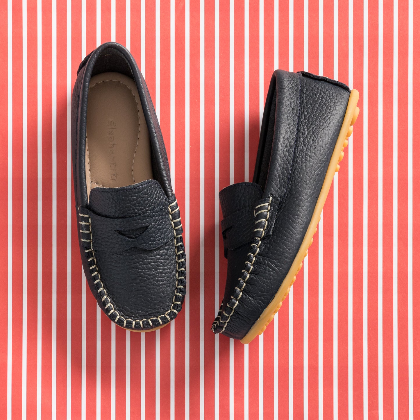Navy blue toddler leather loafers with moccasin stitching and tan rubber soles on red striped background.