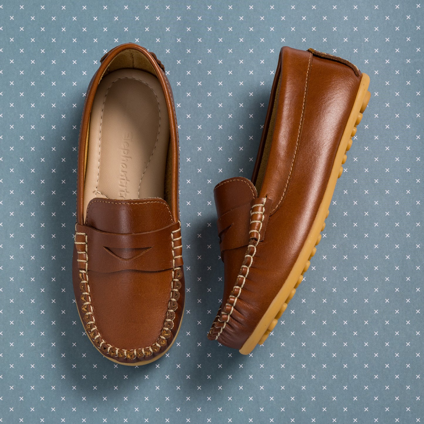 Natural brown leather boys loafers with moccasin stitching and rubber soles on blue patterned background.
