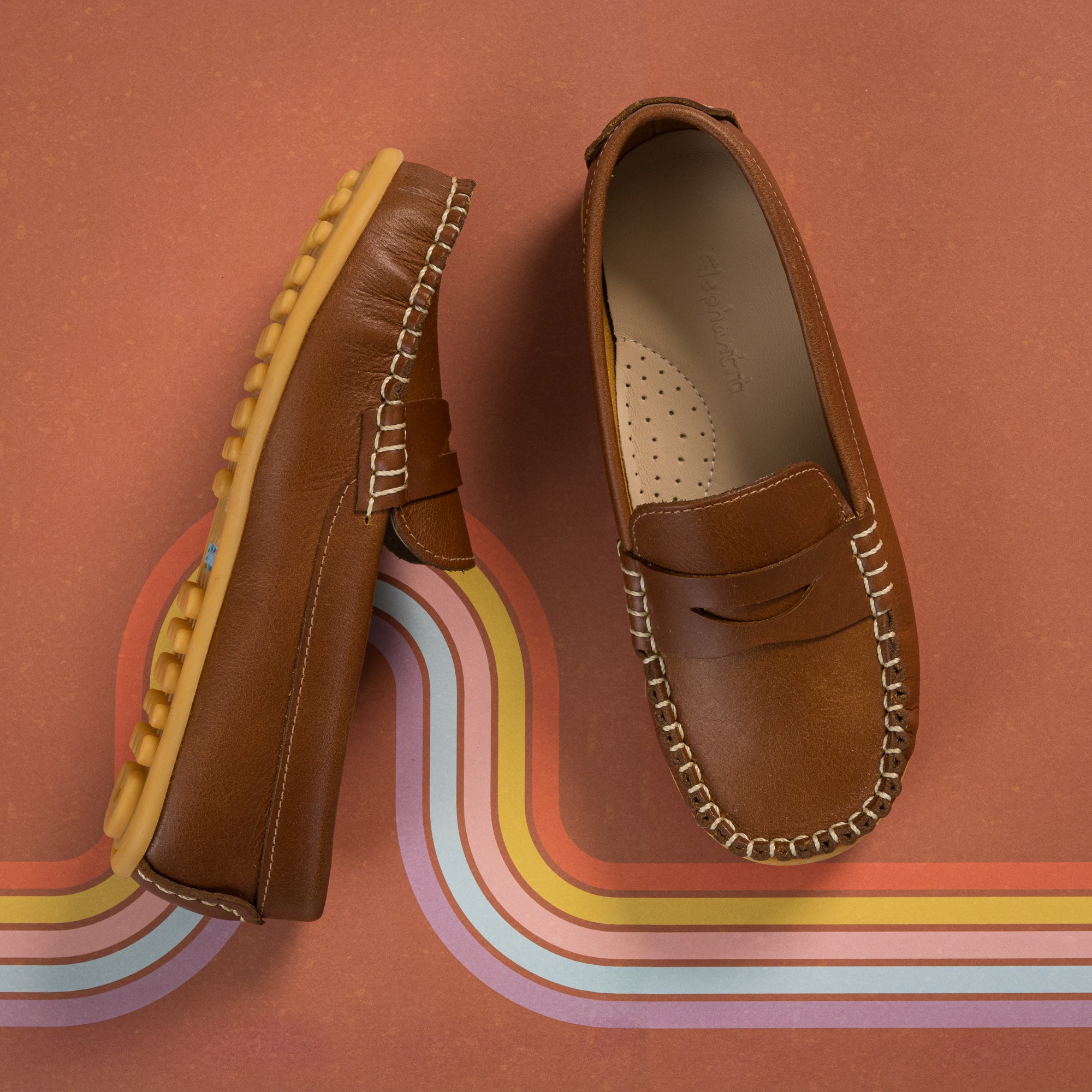Natural brown leather children's loafer with moccasin stitching and rubber sole on a retro striped background.