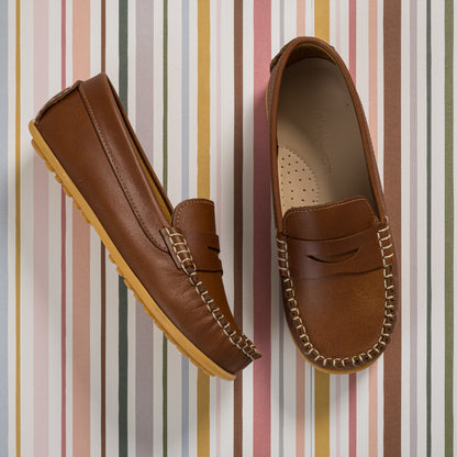 Natural brown leather boys' loafer with moccasin stitching and rubber sole on striped background.