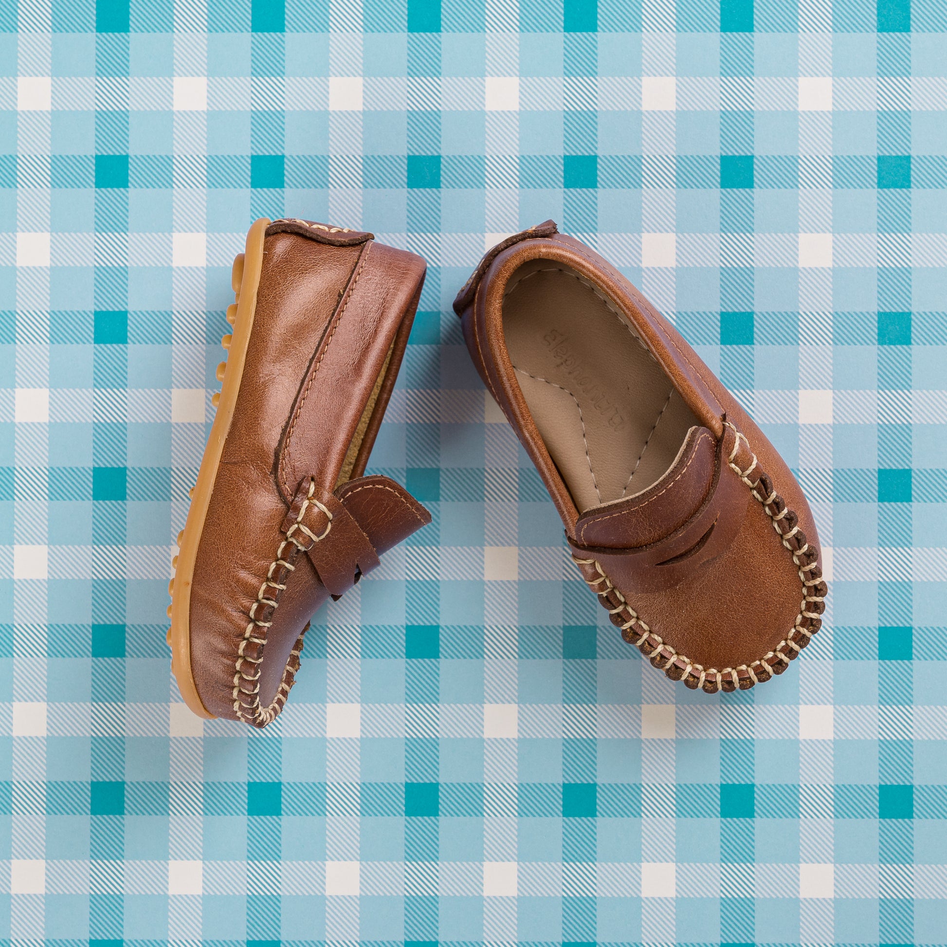 Natural brown leather toddler driving moccasins with stitched detailing and rubber sole on blue plaid background.