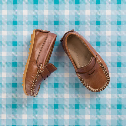 Natural brown leather toddler driving moccasins with stitched detailing and rubber sole on blue plaid background.