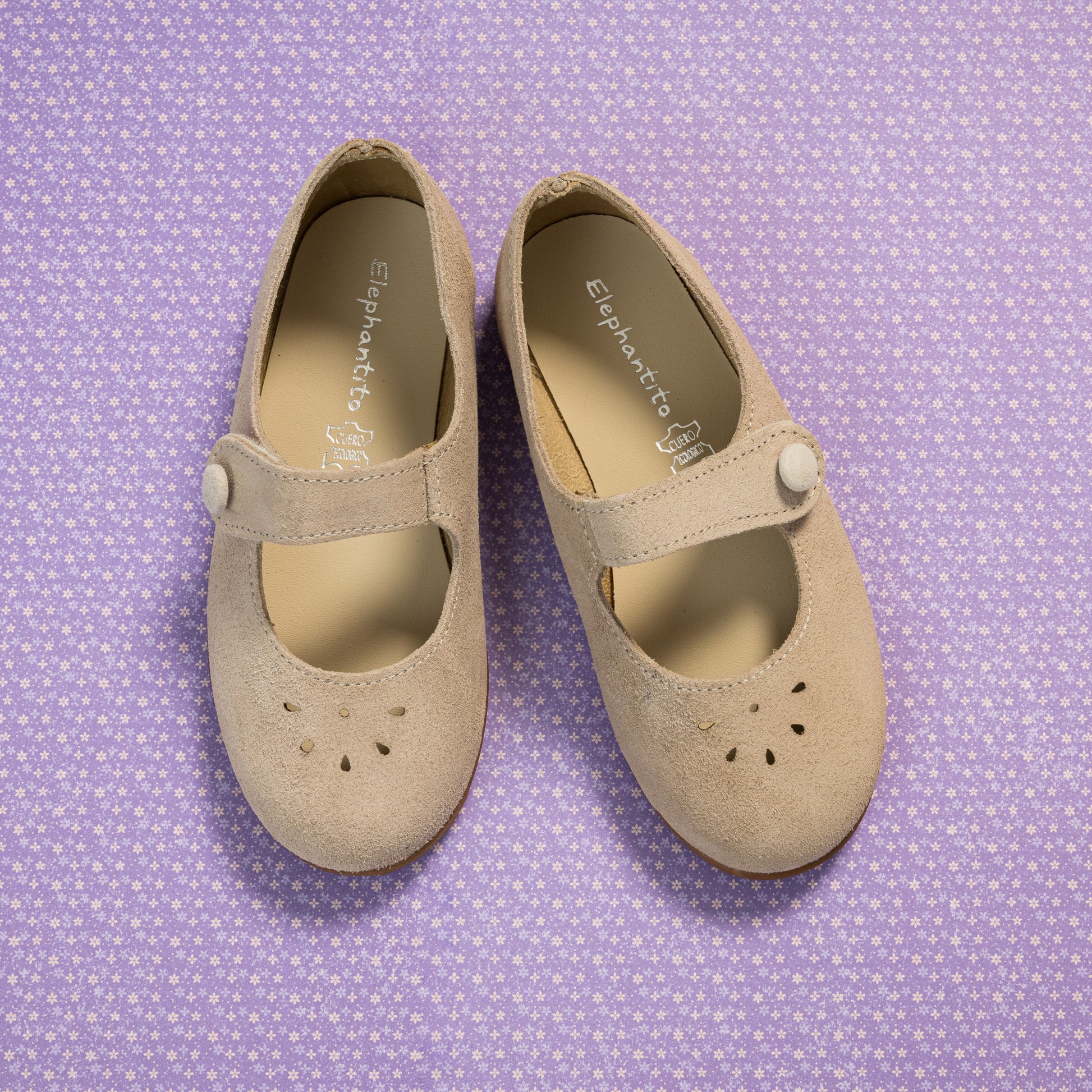 Beige suede Mary Jane shoes for toddlers with button strap and perforated toe design on purple patterned background.