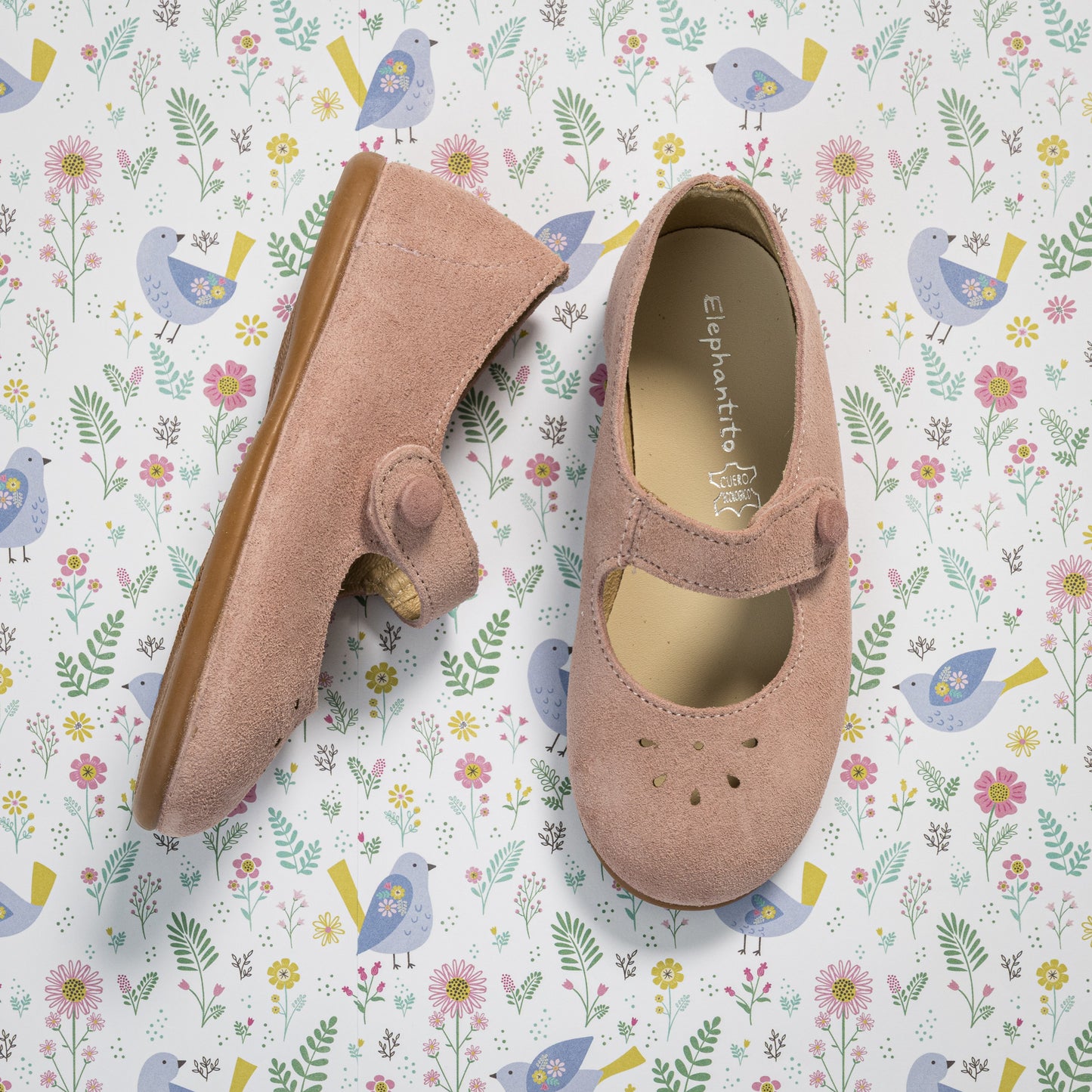 Pink suede toddler Mary Jane shoes with scalloped edges and strap closure on floral background.
