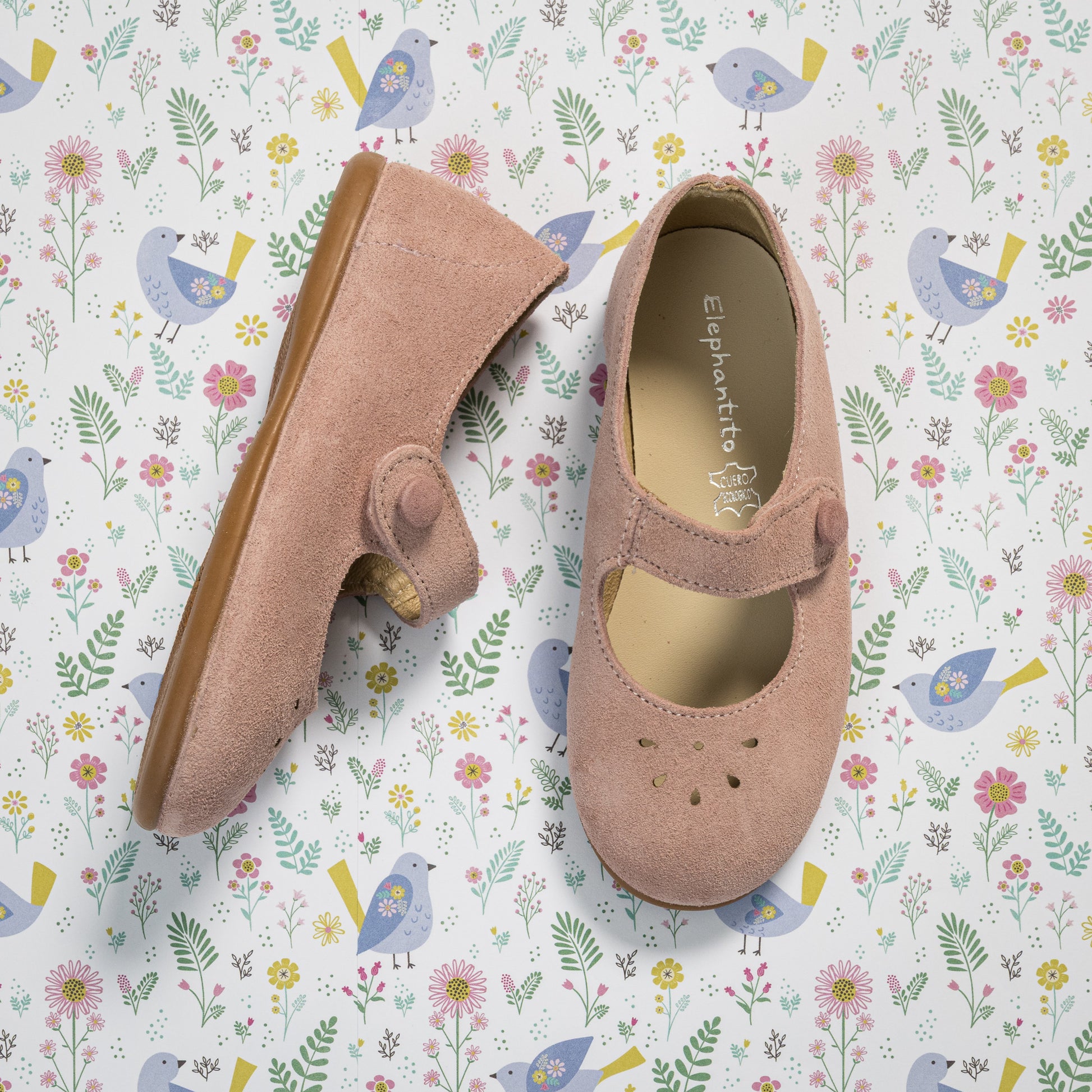 Pink suede toddler Mary Jane shoes with scalloped edges and strap closure on floral background.