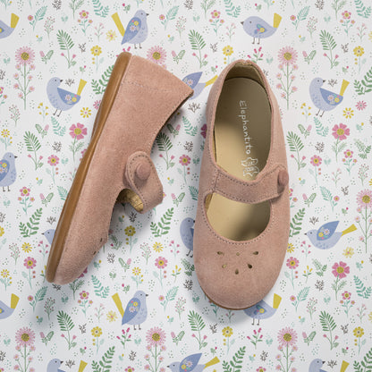 Pink suede toddler Mary Jane shoes with scalloped edges and strap closure on floral background.