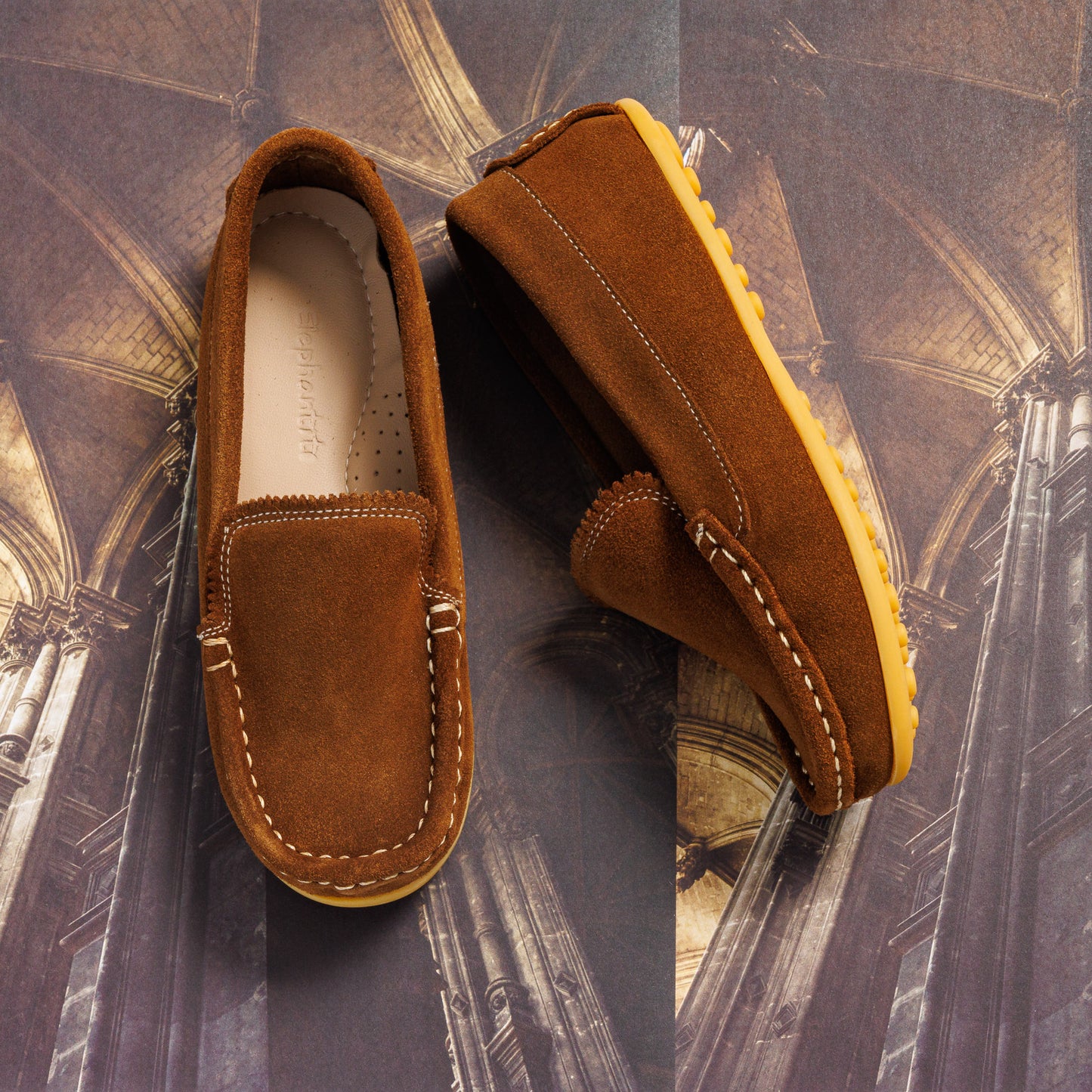Chestnut suede toddler moccasin with white stitching and gum rubber sole on architectural background.