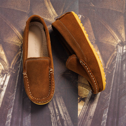 Chestnut suede toddler moccasin with white stitching and gum rubber sole on architectural background.