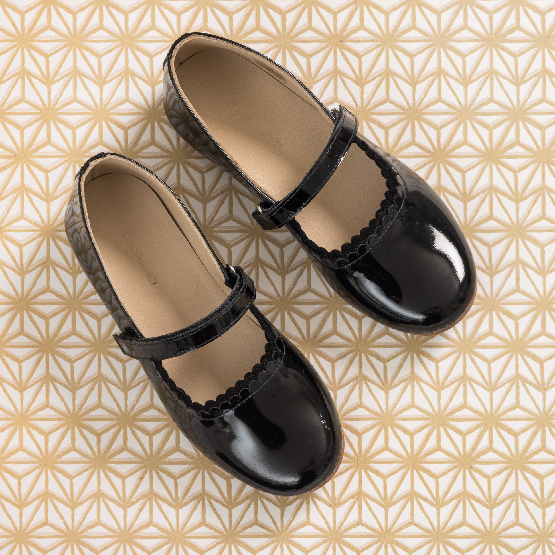 Black patent leather girls' Mary Jane flats with scalloped edges and adjustable strap on geometric patterned background.