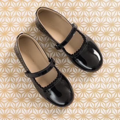 Black patent leather girls' Mary Jane flats with scalloped edges and adjustable strap on geometric patterned background.