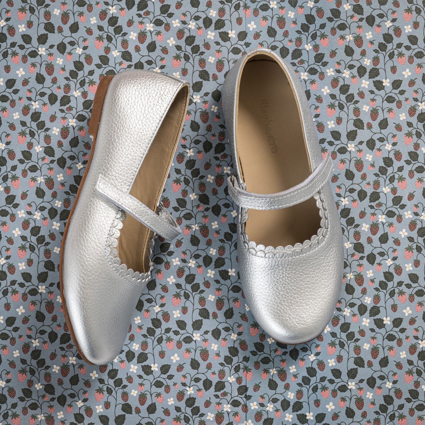 Silver textured leather Mary Jane shoes with scalloped edges and a single strap over the instep on a patterned background.