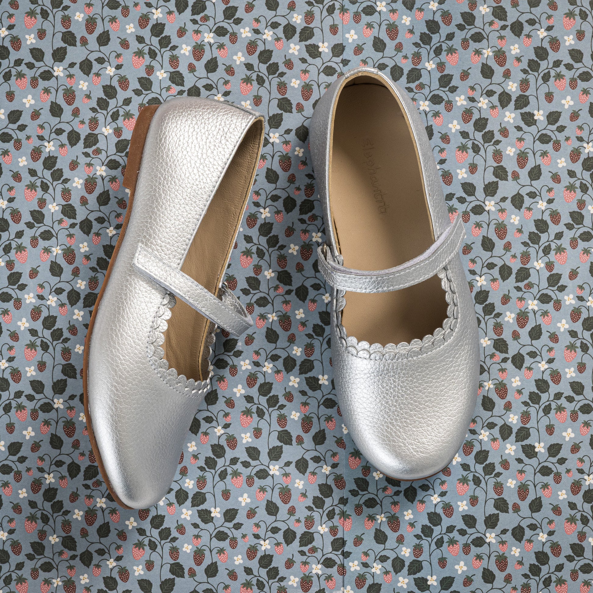 Silver textured leather Mary Jane shoes with scalloped edges and a single strap over the instep on a patterned background.