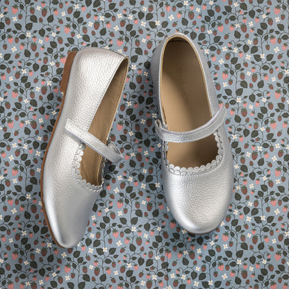 Silver textured leather Mary Jane shoes with scalloped edges and a single strap over the instep on a patterned background.