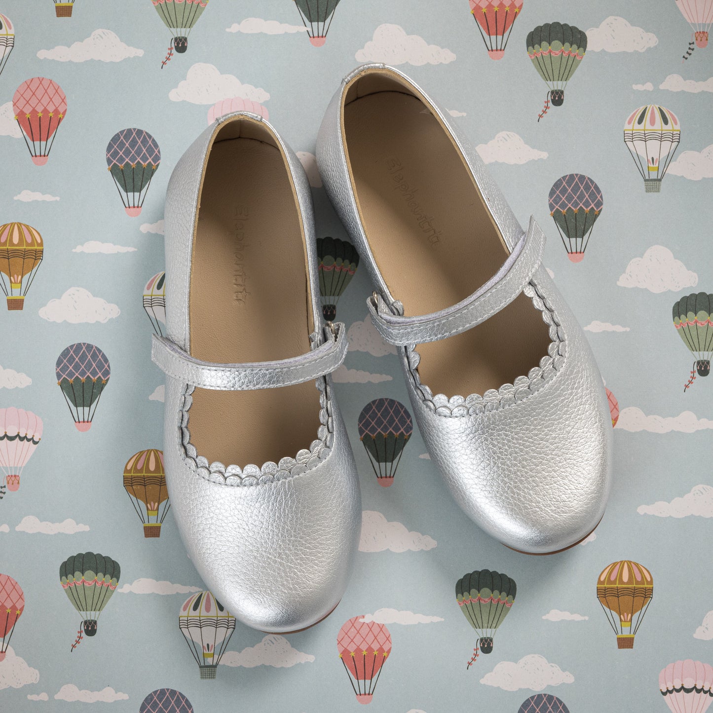 Silver metallic toddler Mary Jane shoes with scalloped edges and Velcro strap on hot air balloon background.