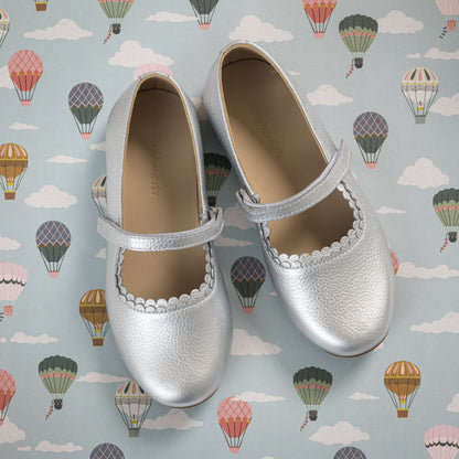 Silver metallic toddler Mary Jane shoes with scalloped edges and Velcro strap on hot air balloon background.