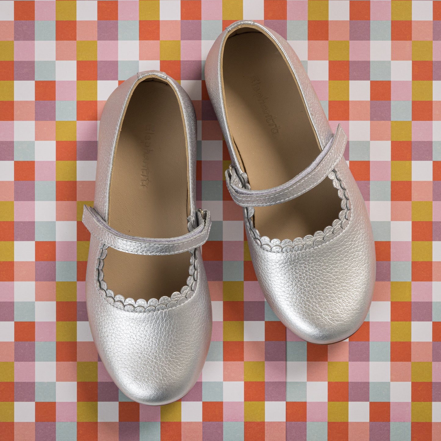 Silver toddler Mary Jane flats with scalloped edges and a Velcro strap on a colorful checkered background.