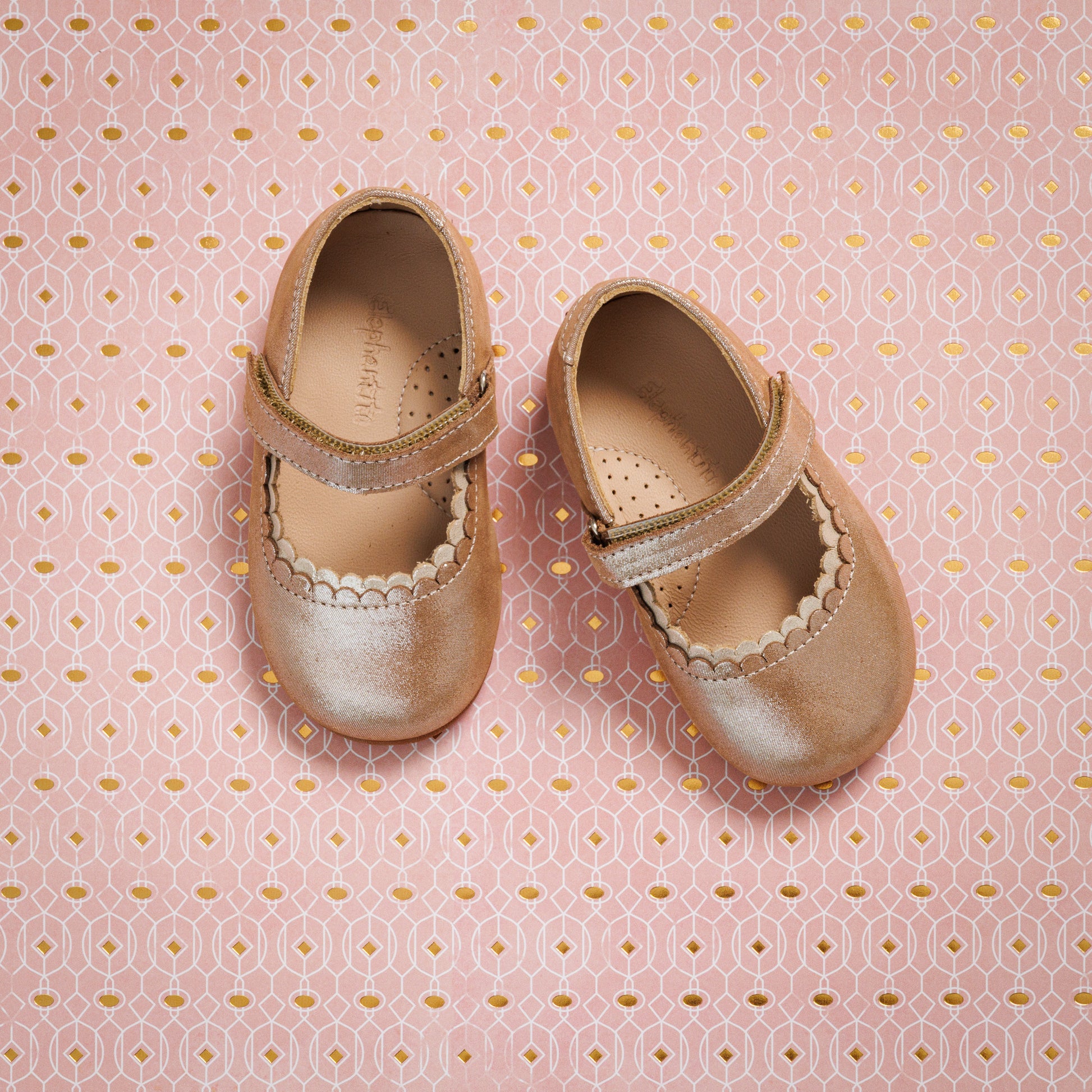 Blush pink toddler Mary Jane shoes with scalloped edges and velcro straps on patterned pink background.