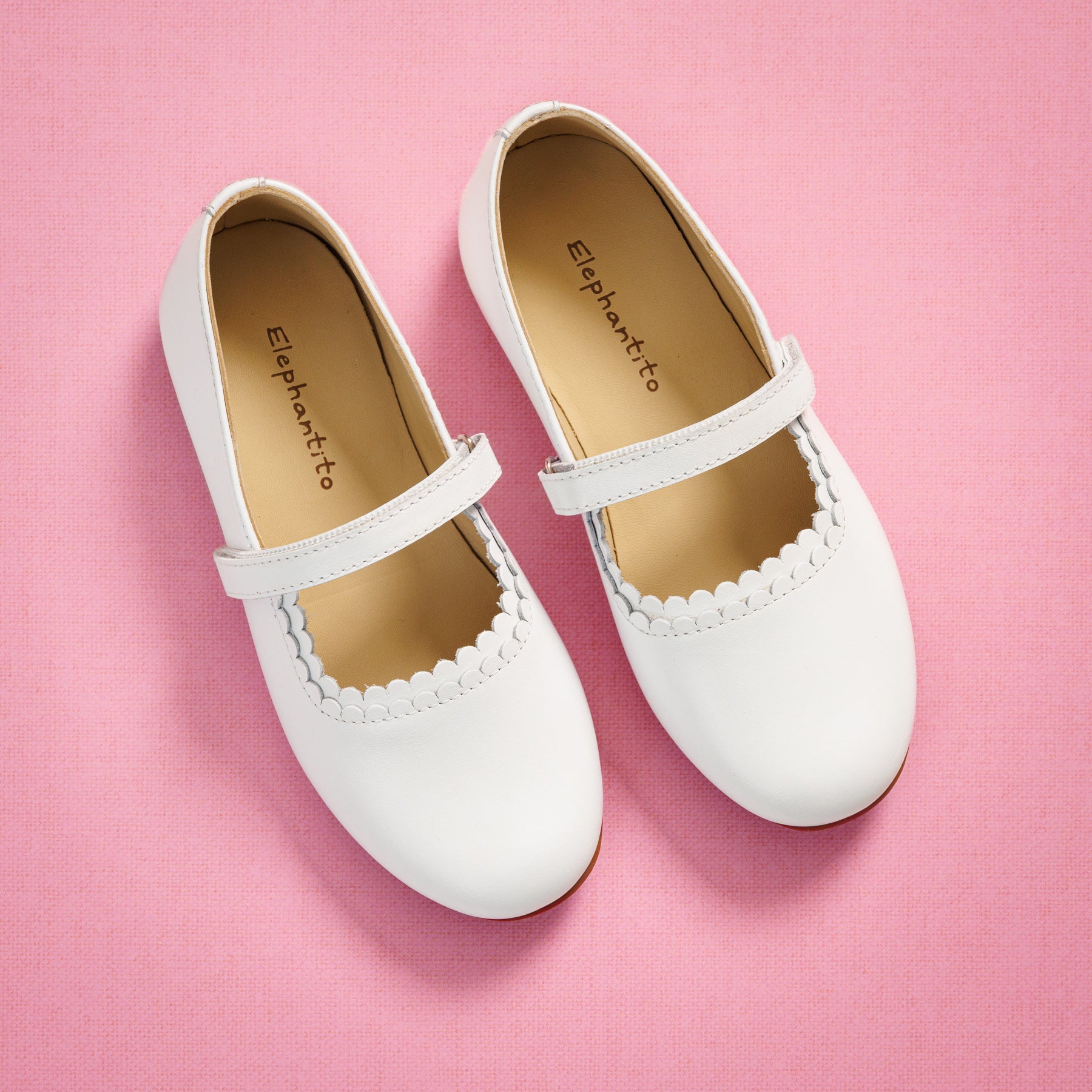 Elephantito - White toddler Mary Jane shoes with scalloped edge and adjustable strap on pink background.