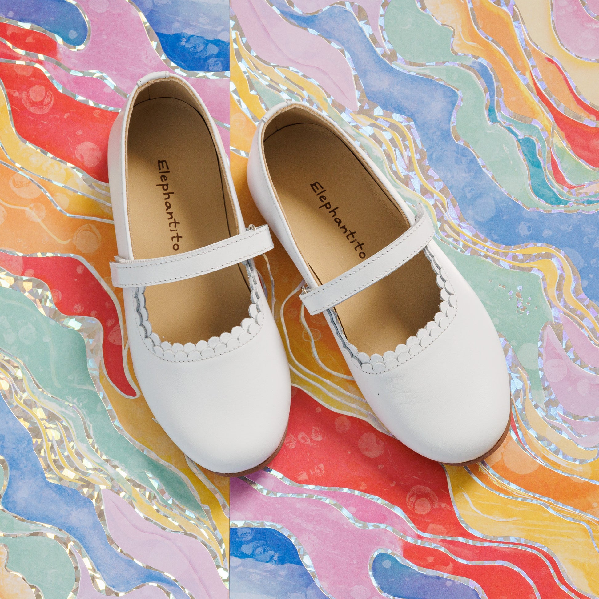 Elephantito - White toddler girls' Mary Jane shoes with scalloped edges and adjustable strap on colorful marbled background.