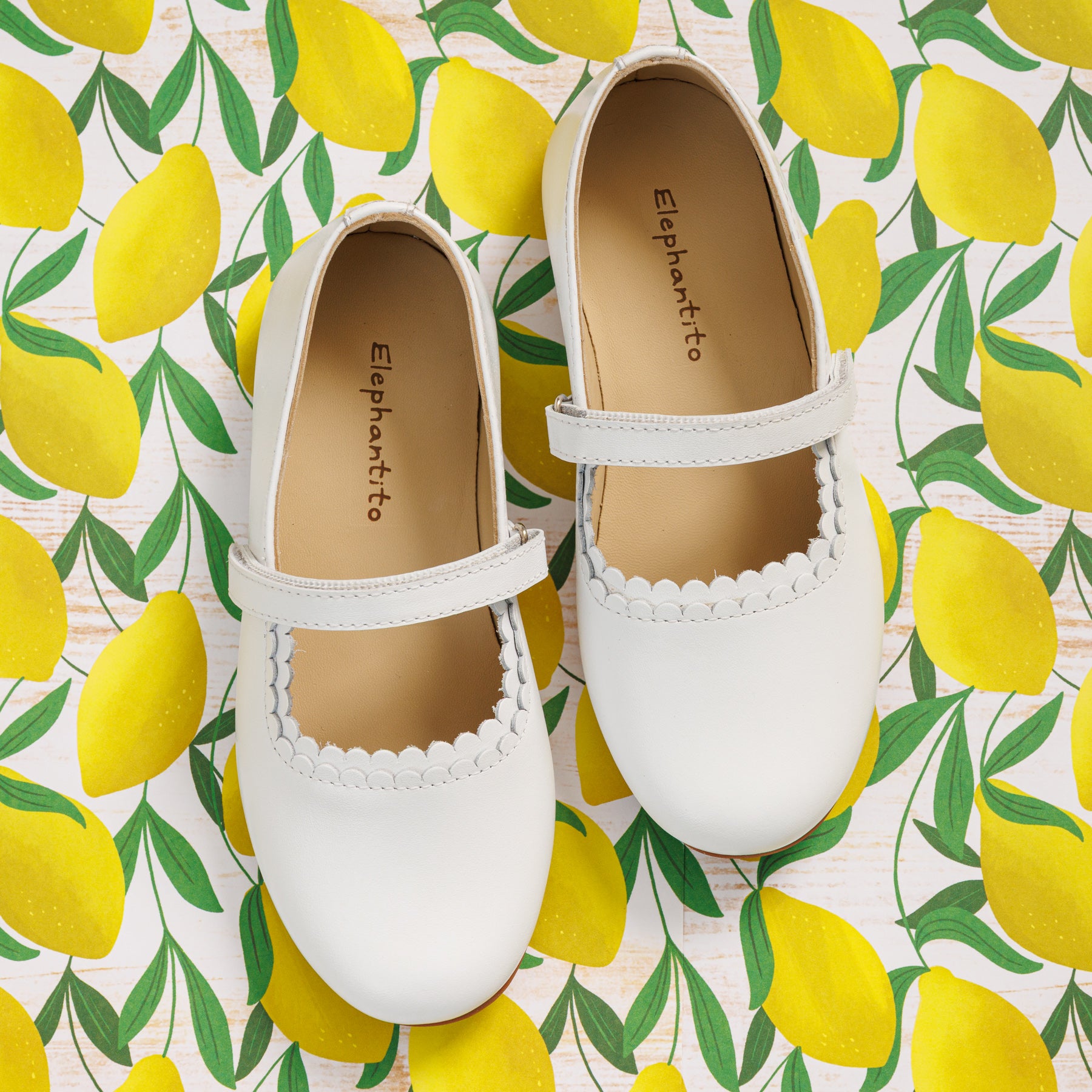Elephantito - White toddler Mary Jane shoes with scalloped edge and Velcro strap on lemon print background.