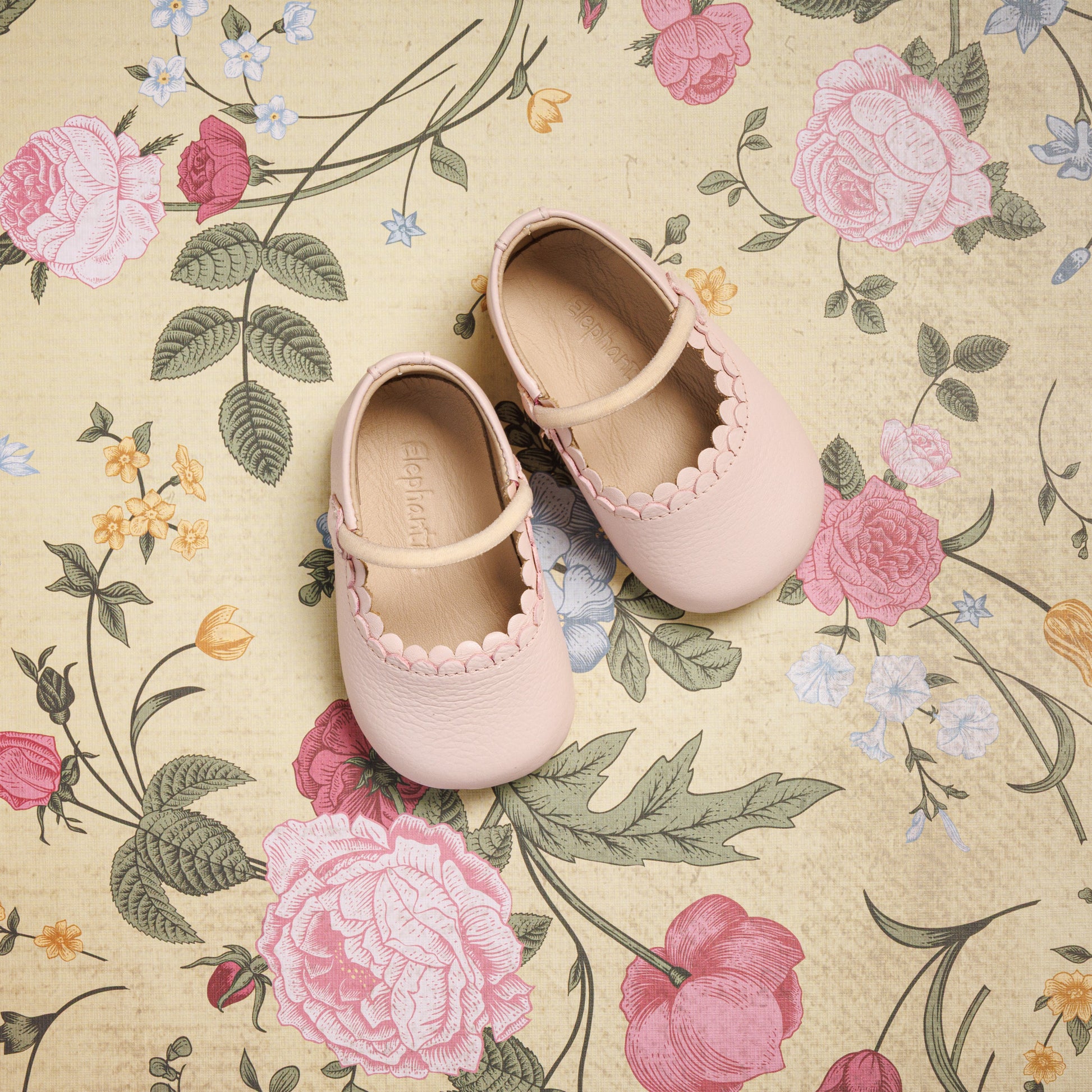 Baby ballet pink Mary Jane shoes with scalloped edges and a single strap on floral background.