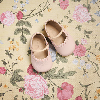 Baby ballet pink Mary Jane shoes with scalloped edges and a single strap on floral background.