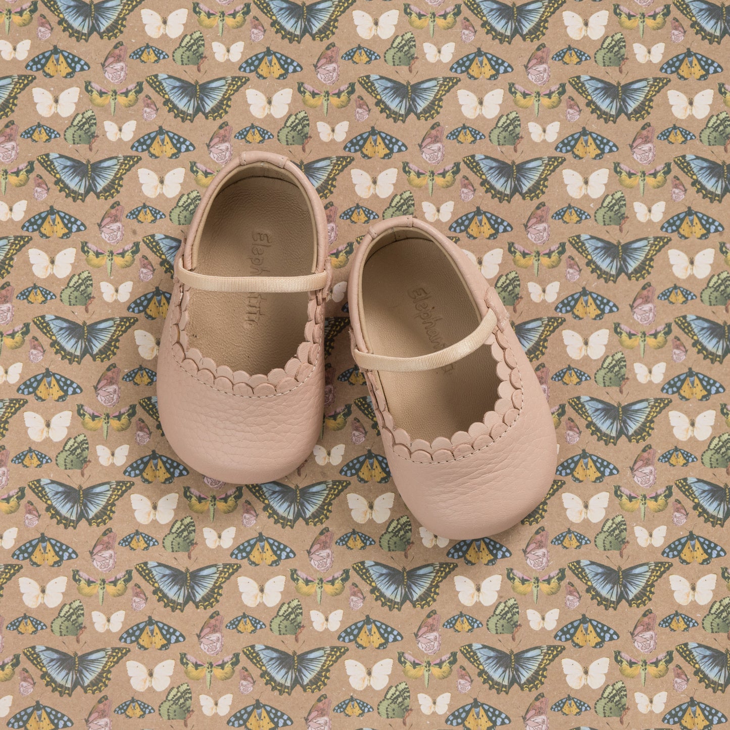Soft pink baby shoes with scalloped edges and a small strap, displayed on patterned fabric with butterflies and flowers.