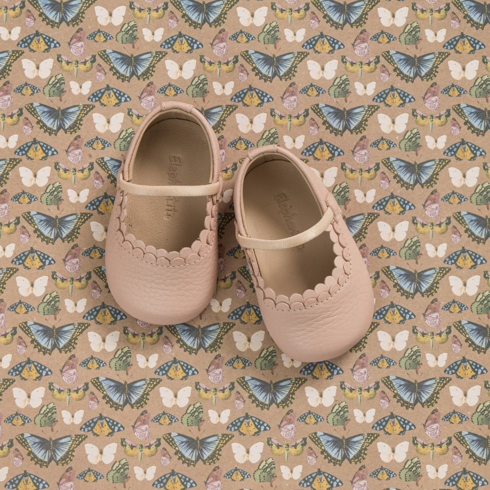 Soft pink baby shoes with scalloped edges and a small strap, displayed on patterned fabric with butterflies and flowers.