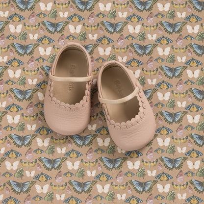 Soft pink baby shoes with scalloped edges and a small strap, displayed on patterned fabric with butterflies and flowers.